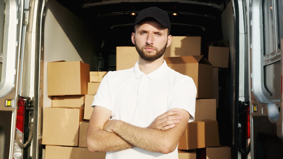 A man wearing a white polo shirt and a black cap stands with arms crossed inside the open rear of a moving van, surrounded by numerous cardboard boxes stacked carefully, some wrapped in plastic. The van is parked on a paved area, with the interior visible showing additional packed boxes and packing materials, indicating an ongoing home relocation process. Dartford Man and Van’s professional removals service is assisting with furniture transport and packing for a house move in Temple Hill DA2 Dartford, with the focus on loading and securing belongings for transportation.