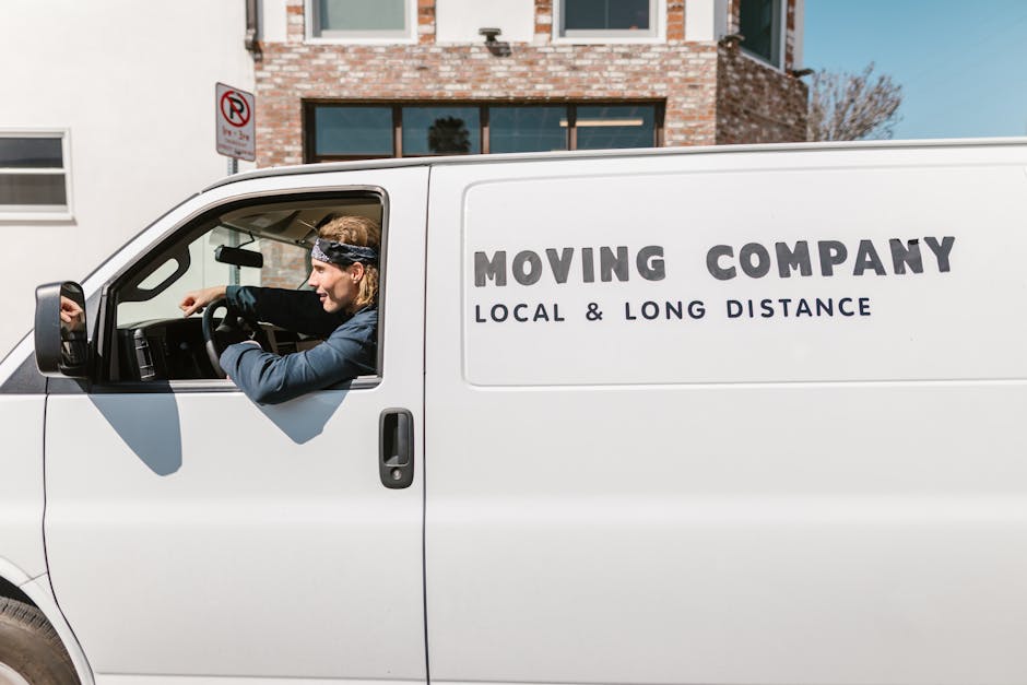 A white moving van parked on a street in front of a brick residential building with large windows and a no-parking sign visible on a nearby pole. A male mover with long hair, wearing a headband and dark clothing, is seated inside the van, leaning out of the open driver’s side window, and appears to be engaged in a conversation or giving instructions. The side of the van displays the company name 'Dartford Man and Van' along with the services of local and long-distance removals, suggesting a house relocation or furniture transport service. The image captures the loading process where the vehicle is ready to assist with home moving logistics, including packing, transporting furniture, and boxes. Bright daylight illuminates the scene, with the mover prepared for a house removal or relocation task.