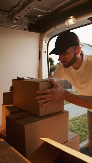 A man wearing a white polo shirt and a black cap stands with arms crossed inside the open rear of a moving van, surrounded by numerous cardboard boxes stacked carefully, some wrapped in plastic. The van is parked on a paved area, with the interior visible showing additional packed boxes and packing materials, indicating an ongoing home relocation process. Dartford Man and Van’s professional removals service is assisting with furniture transport and packing for a house move in Temple Hill DA2 Dartford, with the focus on loading and securing belongings for transportation.