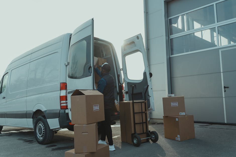 A worker from Dartford Man and Van is loading cardboard boxes and packing materials into the rear of a white cargo van parked outdoors near an industrial building with large glass windows. The van's rear doors are open, revealing a partially packed interior with moving supplies. Several boxes are placed on the pavement beside the vehicle, some stacked and others awaiting transport. The worker, dressed in casual clothing, uses a hand truck to assist with the moving process, indicating a home relocation or furniture transport operation. The scene is illuminated by natural sunlight, suggesting daytime, and captures the logistical aspect of packing and loading during a professional removals service, demonstrating careful handling of packed items as part of house removals near Dartford Train Station, DARTFORD. Dartford Man and Van specializes in removals, supporting clients with efficient house moving solutions.