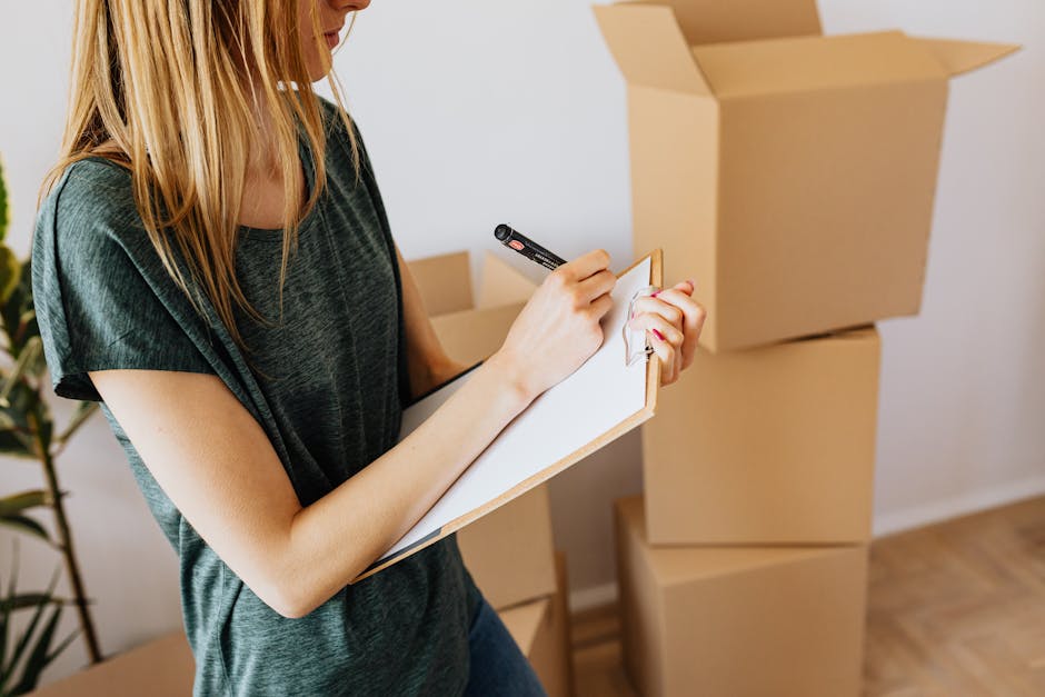 A woman with long blonde hair wearing a dark grey T-shirt is standing indoors, holding a black marker in her right hand and a small whiteboard in her left hand, which she is writing on. Behind her, there are three large, open-top cardboard boxes stacked on the floor, with another box positioned on top of the stack. The boxes are light brown, made of corrugated cardboard, and are ready for packing or transport within a home relocation process. To the left, there's a glimpse of a green potted plant with long, pointed leaves, suggesting a domestic environment. The scene is well-lit with natural light, indicating daytime, and appears to be part of a moving or packing activity associated with house removals, as handled by Dartford Man and Van. The image depicts the packing stage of furniture transport or moving logistics, aligned with professional removal services for relocating belongings from one address to another, such as from Bluewater to Dartford.