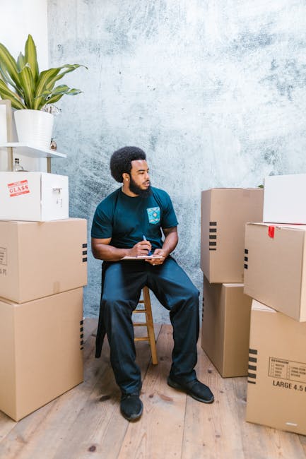 A man with dark skin, a full beard, and short curly hair, wearing a blue company uniform, is seated on a wooden chair in a room with a textured light blue wall and wooden flooring. He is surrounded by several large cardboard boxes, some sealed with packing tape, and one box with red tape. The boxes vary in size and are stacked on the floor and against the wall, with some placed on the left and others on the right side of the man. There is a white shelving unit on the left side of the image, holding a potted plant with large green leaves, a white container, and a cardboard box labeled 'GLASS.' The room appears to be set up for packing or organizing items as part of a home relocation process. The man is holding a clipboard and pen, appearing to be taking notes or planning the move, which reflects the packing and moving activities involved in professional removals services offered by Dartford Man and Van. The environment looks well-lit, clean, and organized, suitable for house removals from Bluewater to Dartford.