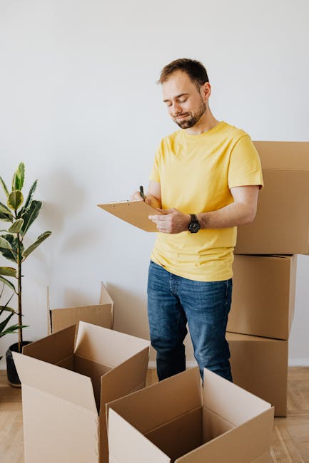 A woman with long blonde hair wearing a dark grey T-shirt is standing indoors, holding a black marker in her right hand and a small whiteboard in her left hand, which she is writing on. Behind her, there are three large, open-top cardboard boxes stacked on the floor, with another box positioned on top of the stack. The boxes are light brown, made of corrugated cardboard, and are ready for packing or transport within a home relocation process. To the left, there's a glimpse of a green potted plant with long, pointed leaves, suggesting a domestic environment. The scene is well-lit with natural light, indicating daytime, and appears to be part of a moving or packing activity associated with house removals, as handled by Dartford Man and Van. The image depicts the packing stage of furniture transport or moving logistics, aligned with professional removal services for relocating belongings from one address to another, such as from Bluewater to Dartford.