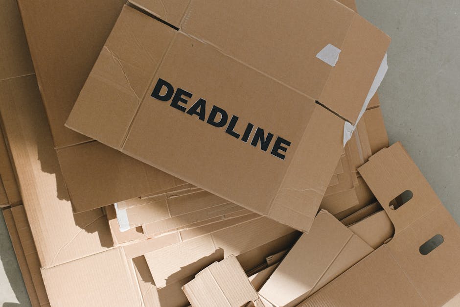 A collection of flattened cardboard boxes, some with handles and cutouts, stacked on top of each other on a floor, with one prominent box displaying the word 'DEADLINE' in bold black letters. The boxe